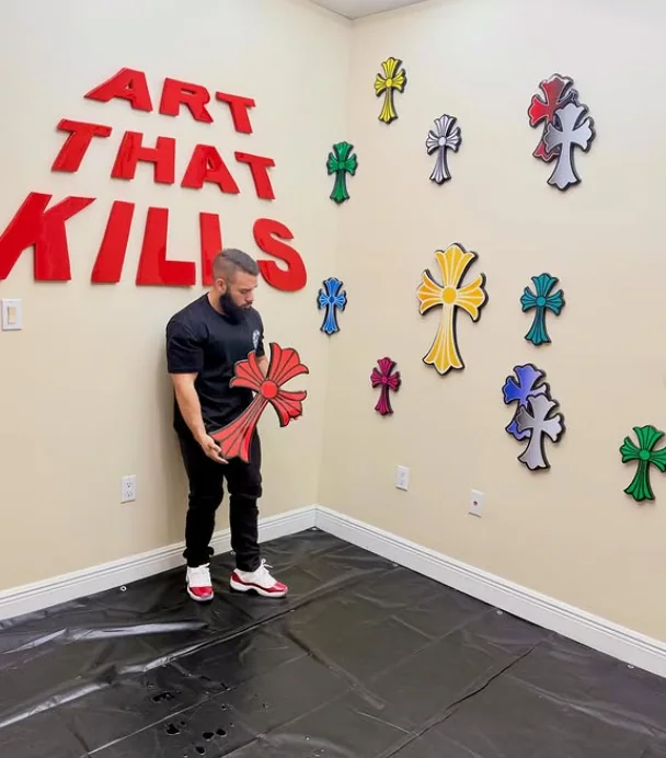Frankie in his studio holding a red epoxy cross, below the words ART THAT KILLS in red lettering, with a wall of coloured crosses beside him